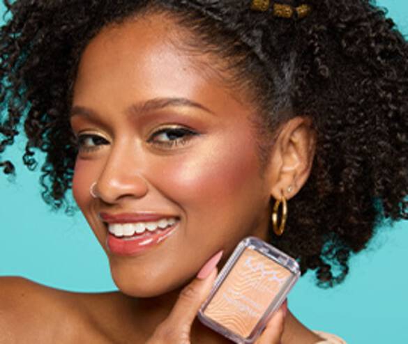 A smiling woman with curly hair holds a blush compact, showcasing her radiant makeup against a bright turquoise background.